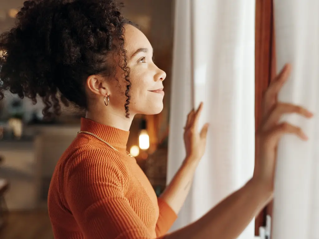 Femme apaisée ouvrant les rideaux de sa fenêtre dans un intérieur chaleureux et lumineux